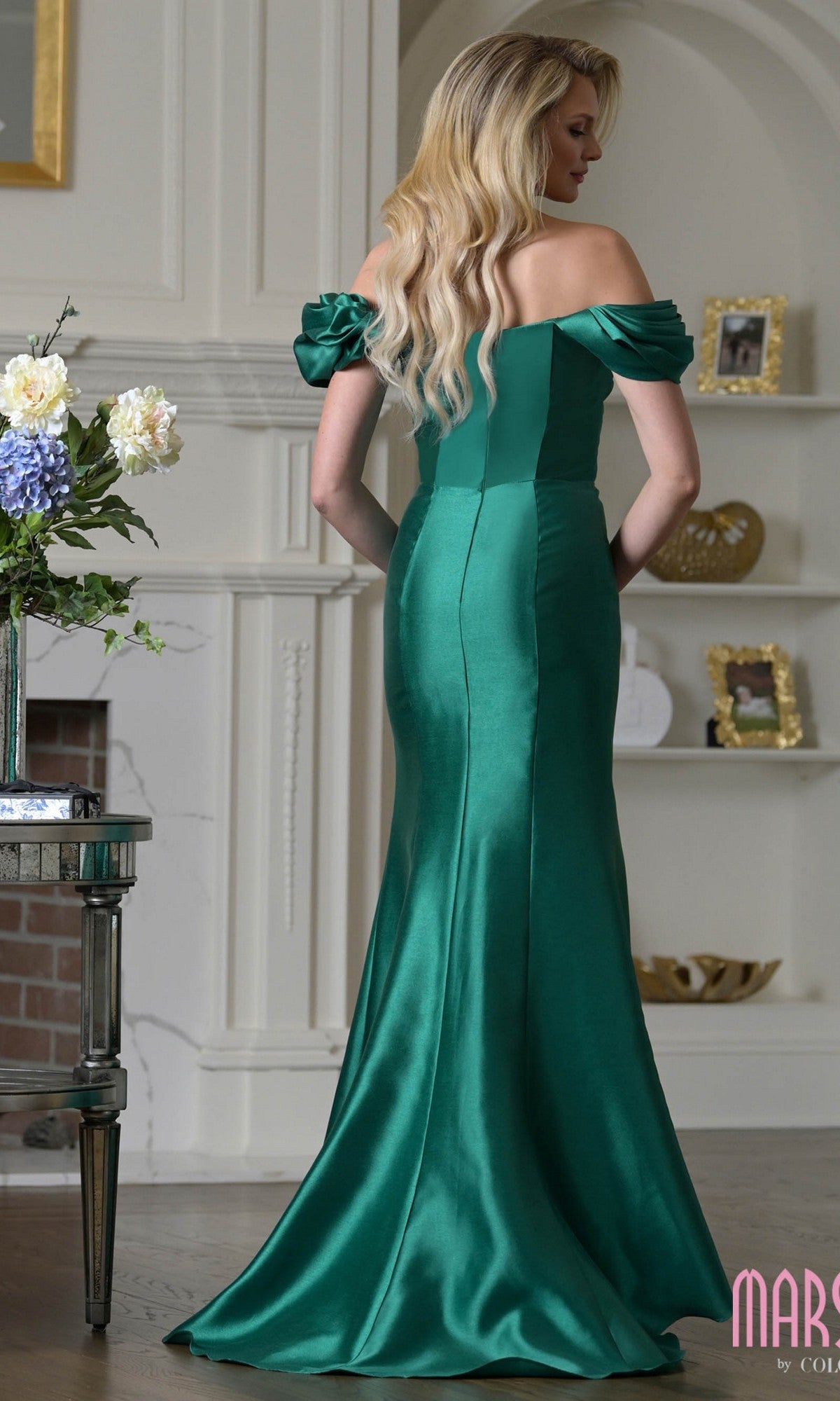 A woman stands indoors in the Marsoni MV1407 Long Formal Dress—an emerald green satin, ruffled off-shoulder, floor-length gown with a fitted bodice, flared skirt, and elegant train. She faces away to highlight the dress’s stunning back details.